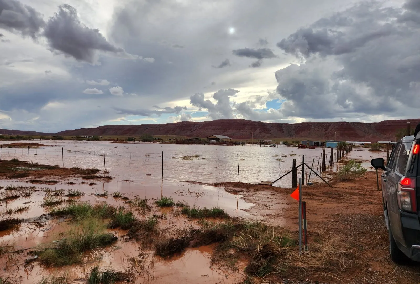 Floodwater covers a rural desert field beneath storm clouds, with fences, buildings, and an SUV visible.