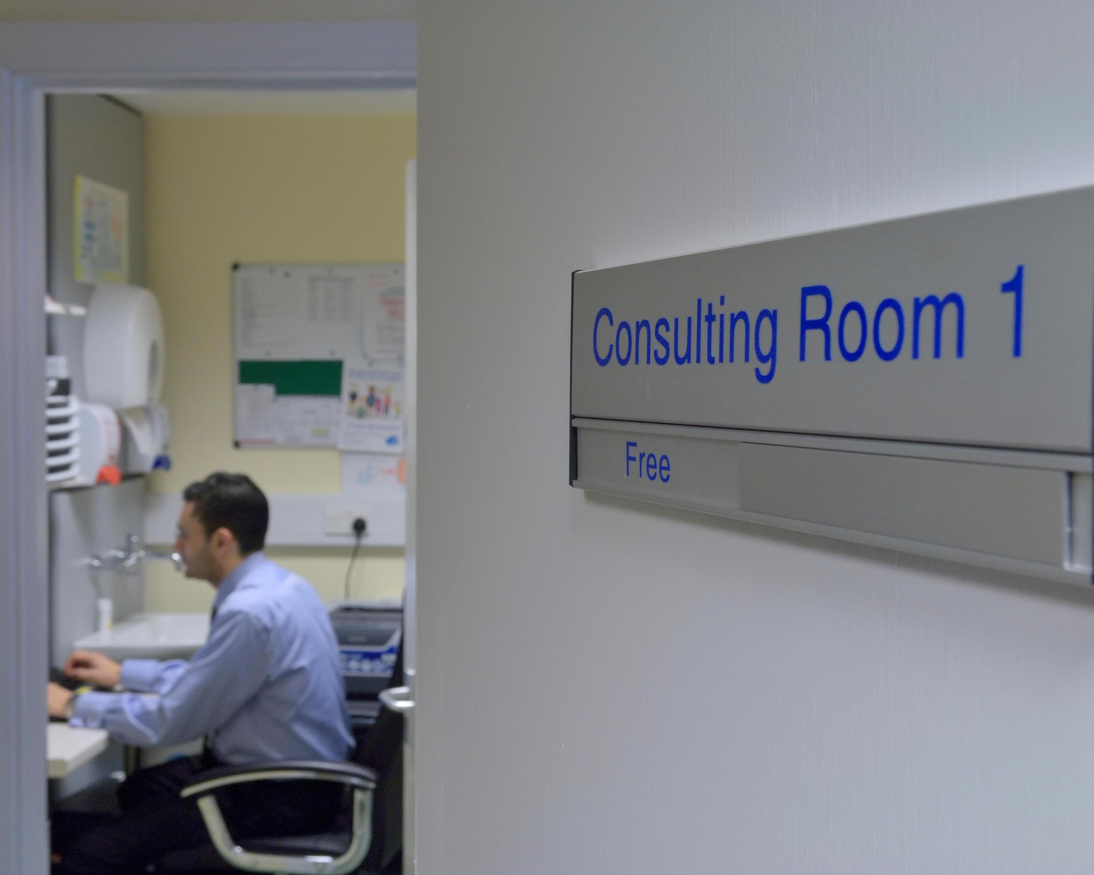 A seated man works at a desk inside a clinic room, viewed past a sign reading “Consulting Room 1.”