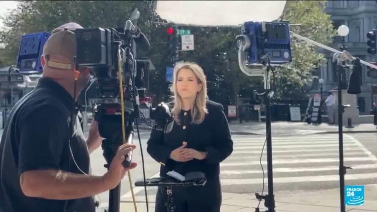 A female reporter stands beside a camera operator and broadcast equipment at a city intersection.