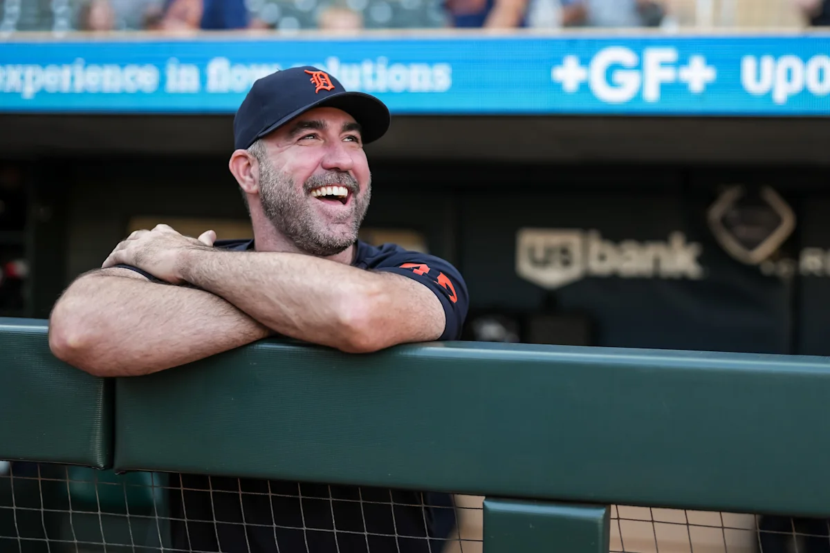 Smiling baseball player in Tigers cap leans on dugout railing with arms crossed.
