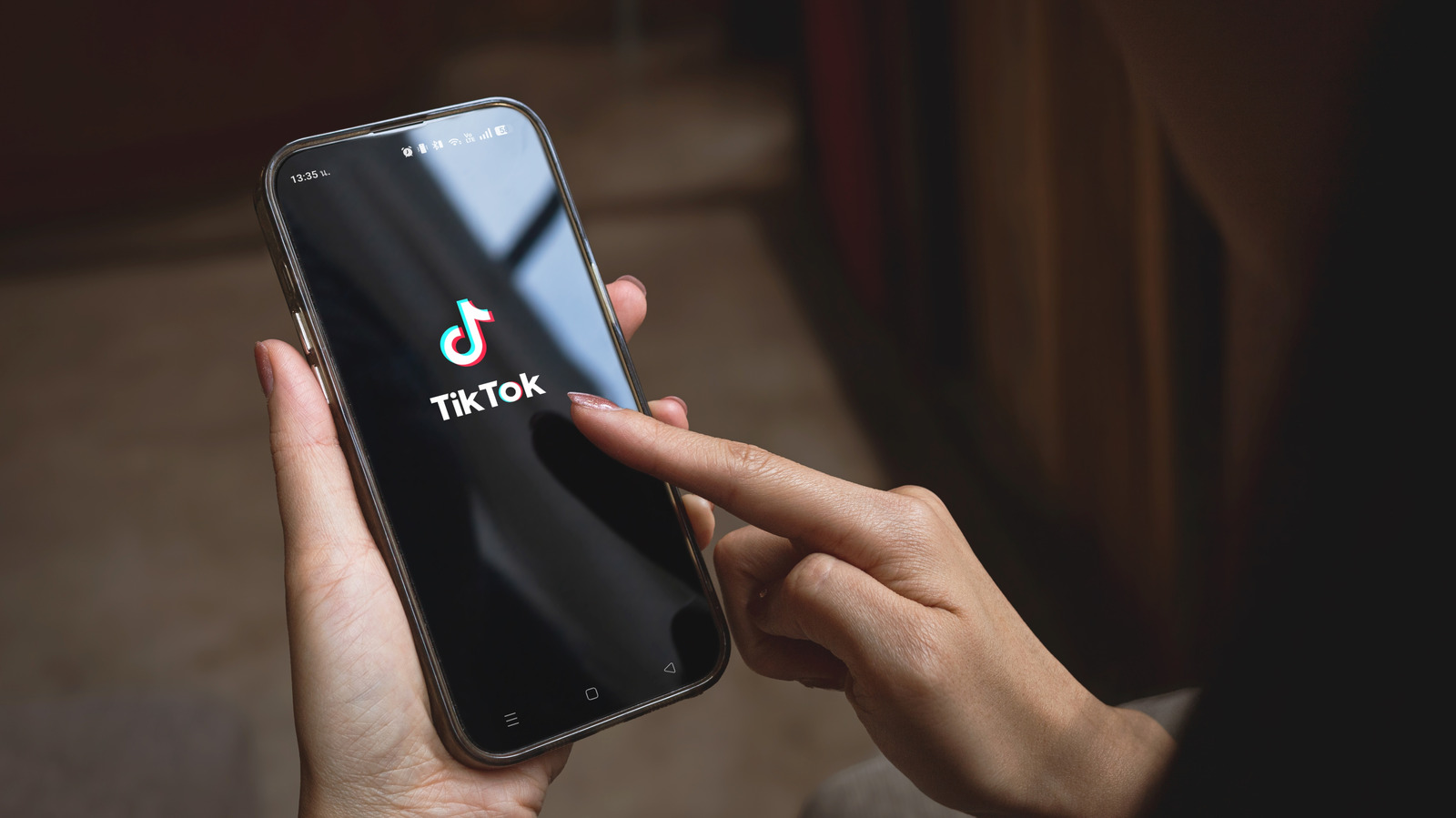 A person holds a smartphone displaying the TikTok logo and taps the screen.