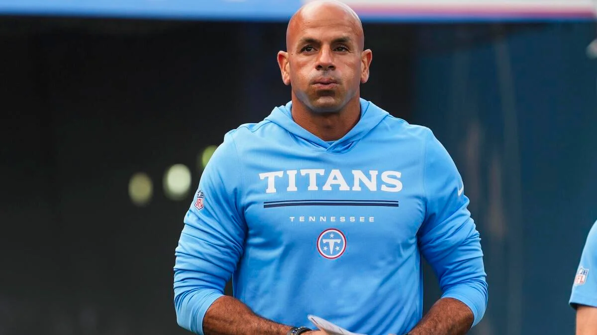 Bald man wearing a light blue Titans hoodie stands on the sideline during a football game.