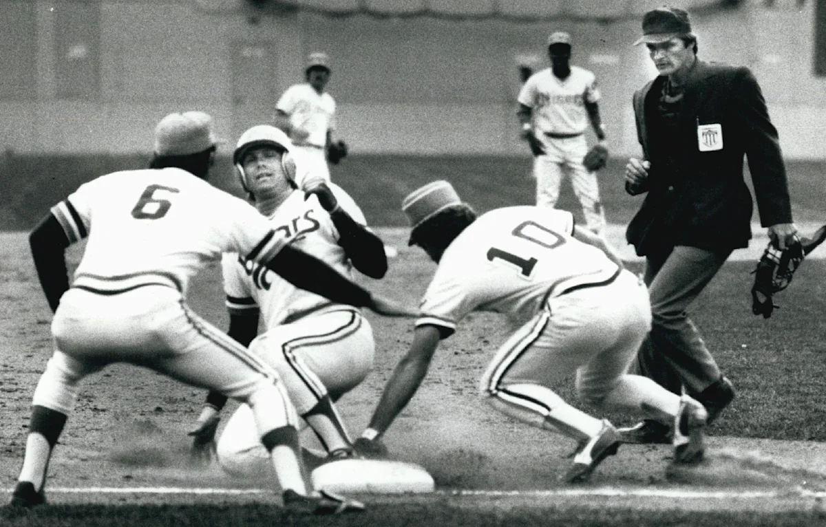 Black-and-white baseball scene with runner sliding as two defenders converge near a base