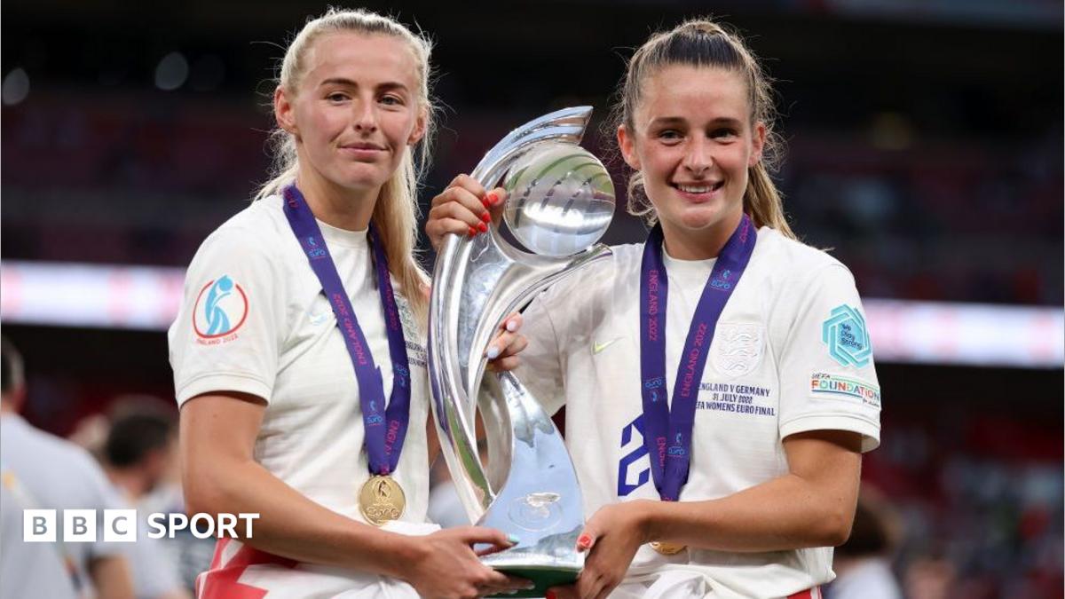 Two England players pose with a silver trophy and medals, wearing white shirts on a stadium pitch.