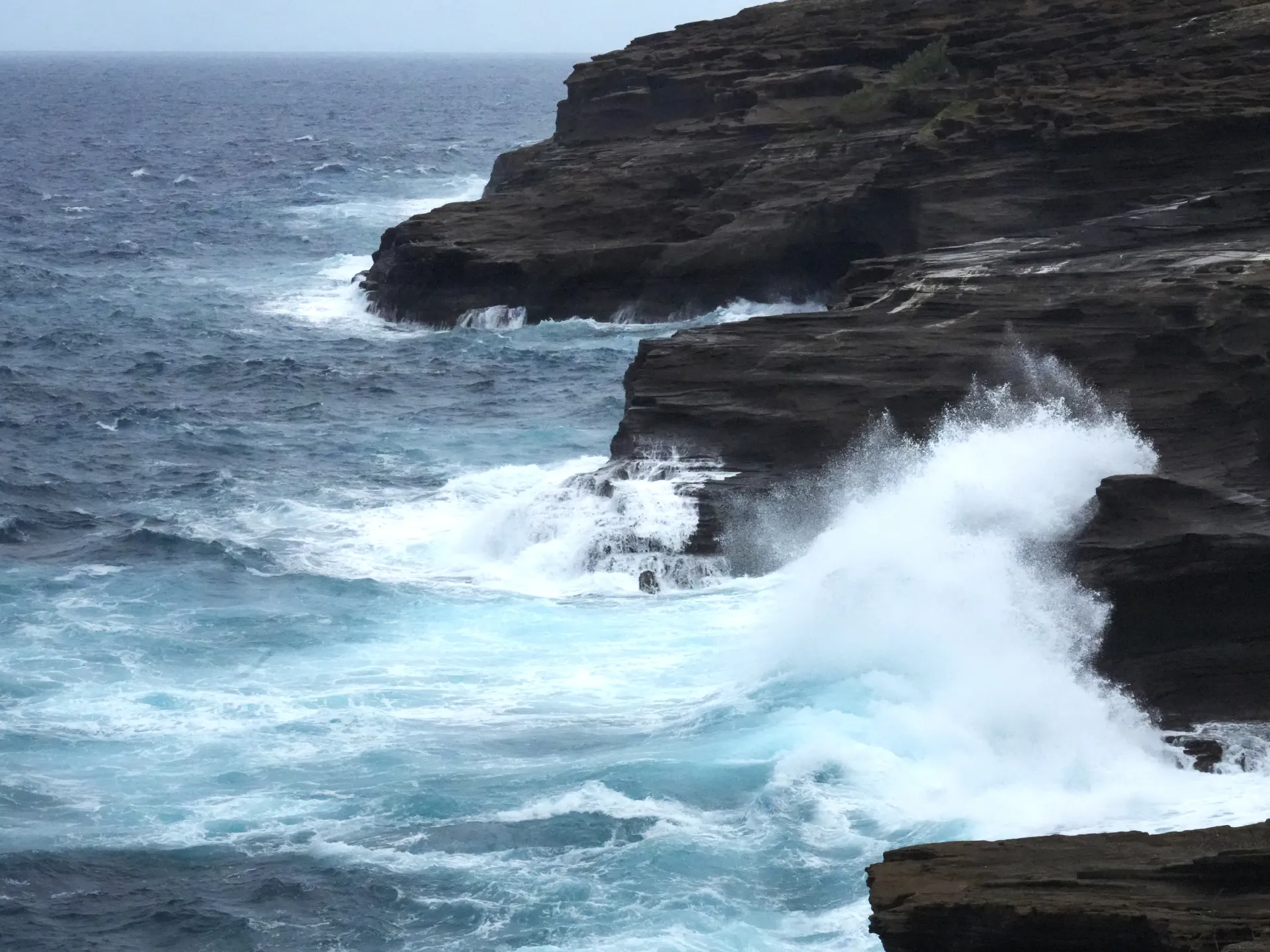 Large waves crash against dark layered coastal cliffs beneath an overcast sky.