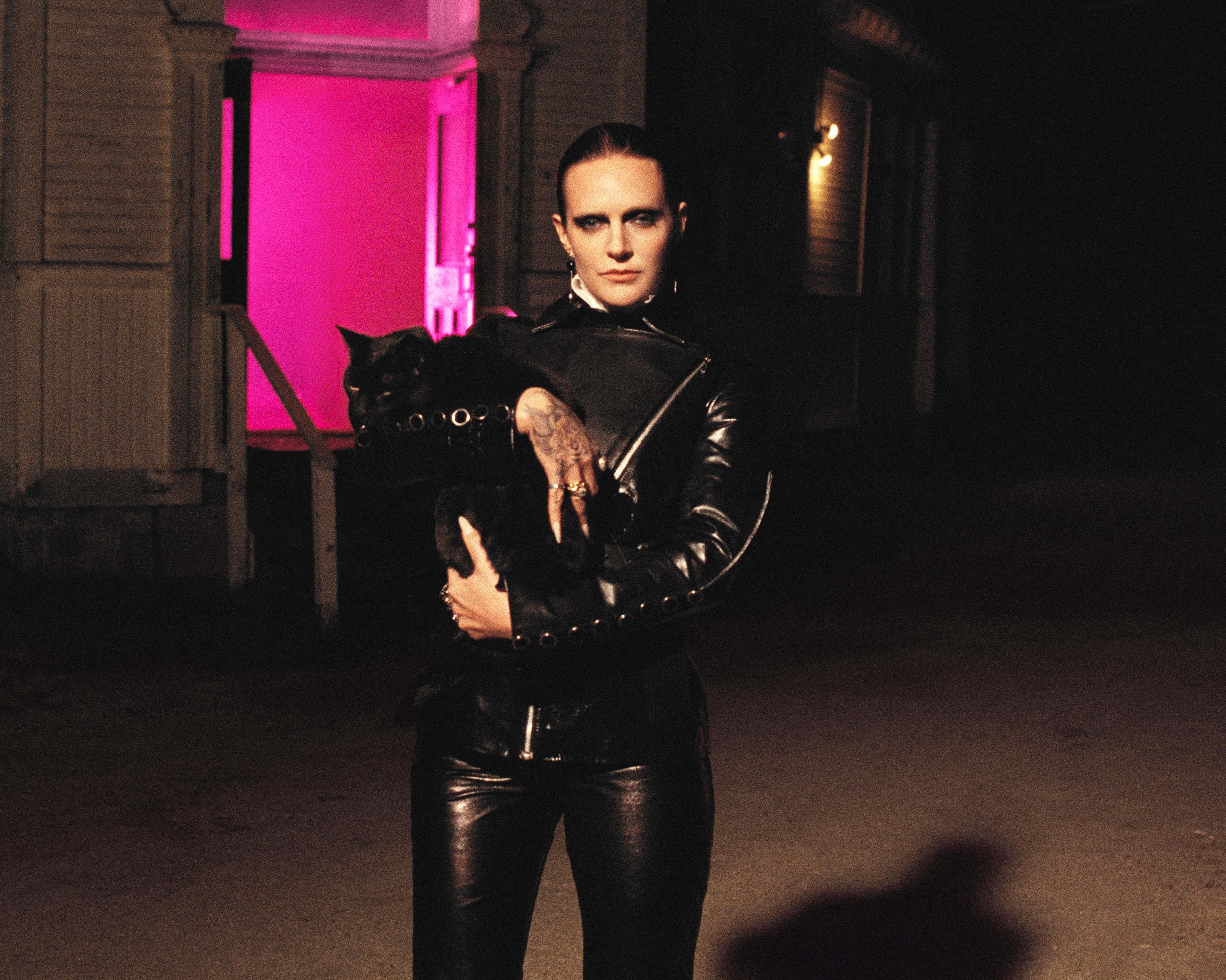 Tattooed woman in black leather holds a black cat outside a dimly lit building.