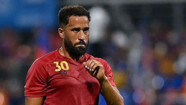 Bearded footballer in a red jersey walks across the pitch during a match.