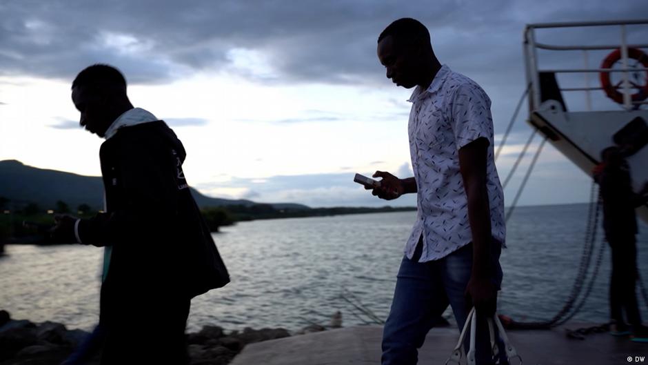 Two men walk beside a waterfront near a boat at dusk, carrying personal belongings.