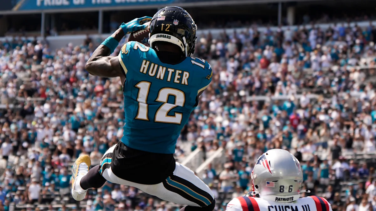 Jaguars player in teal jersey leaps with the ball as a Patriots player watches below.