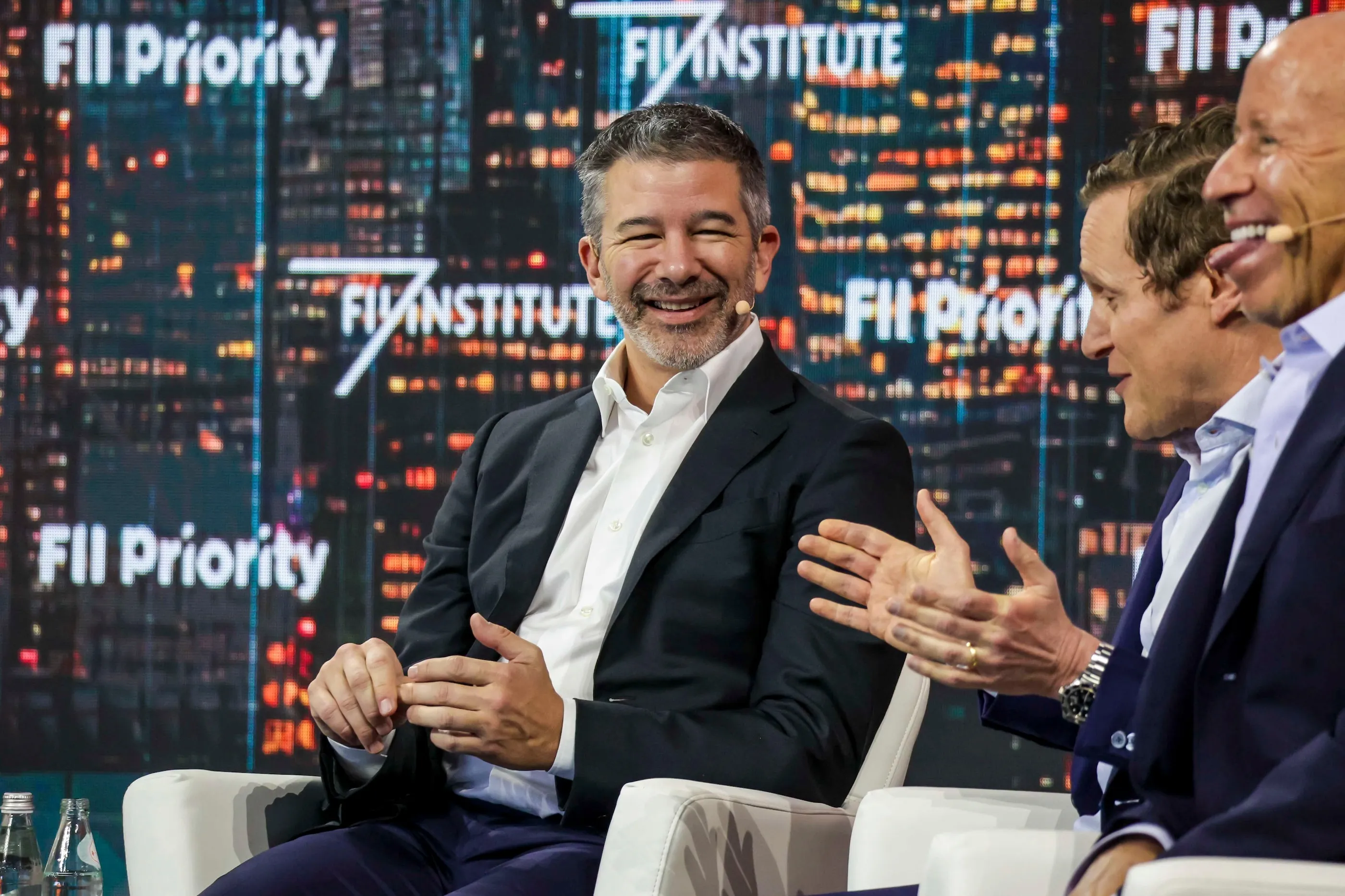 Three men in suits sit onstage during a panel discussion, with technology-themed screens behind them.