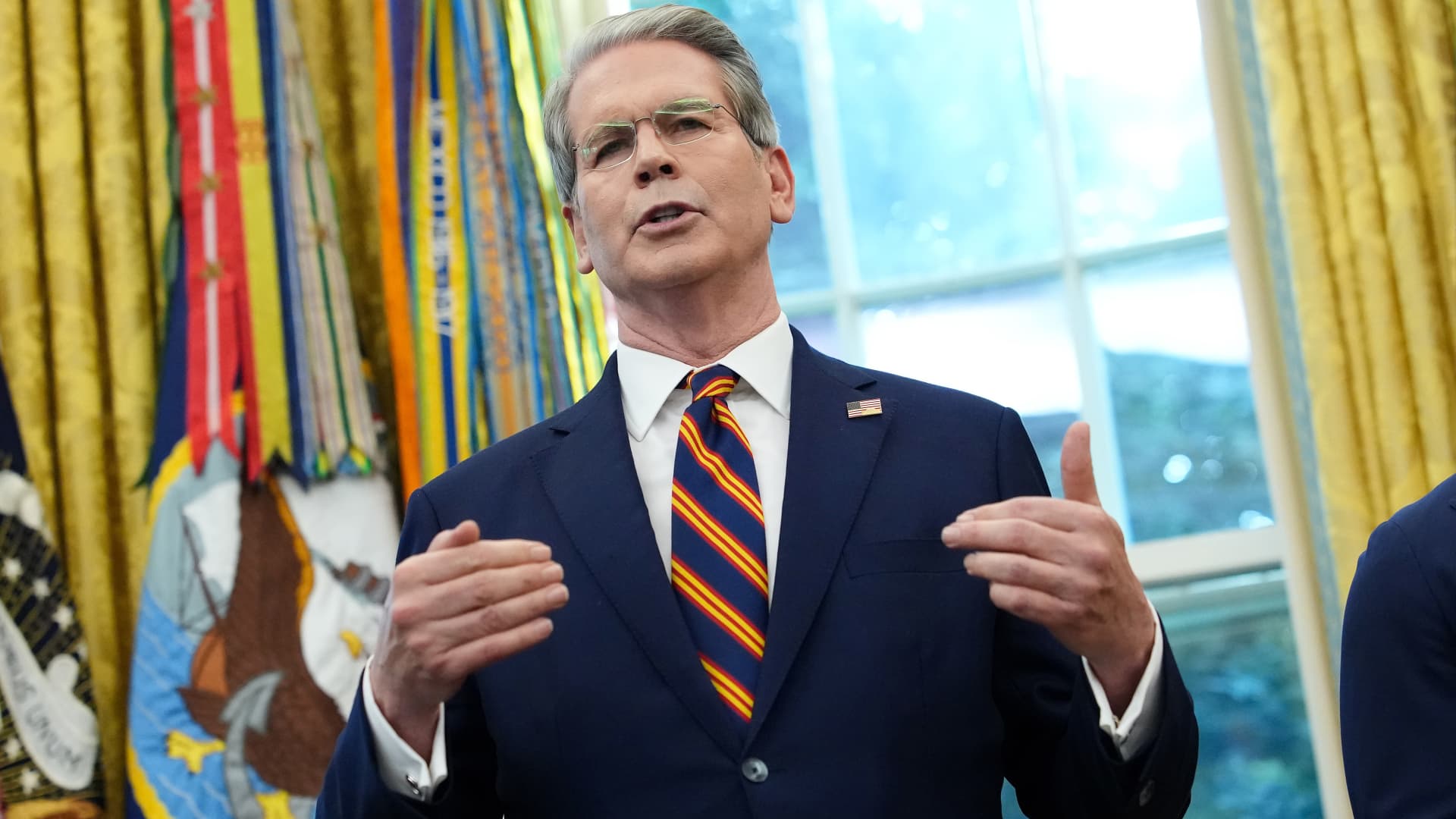 Gray-haired man in navy suit and striped tie gestures while speaking indoors near military ribbons.