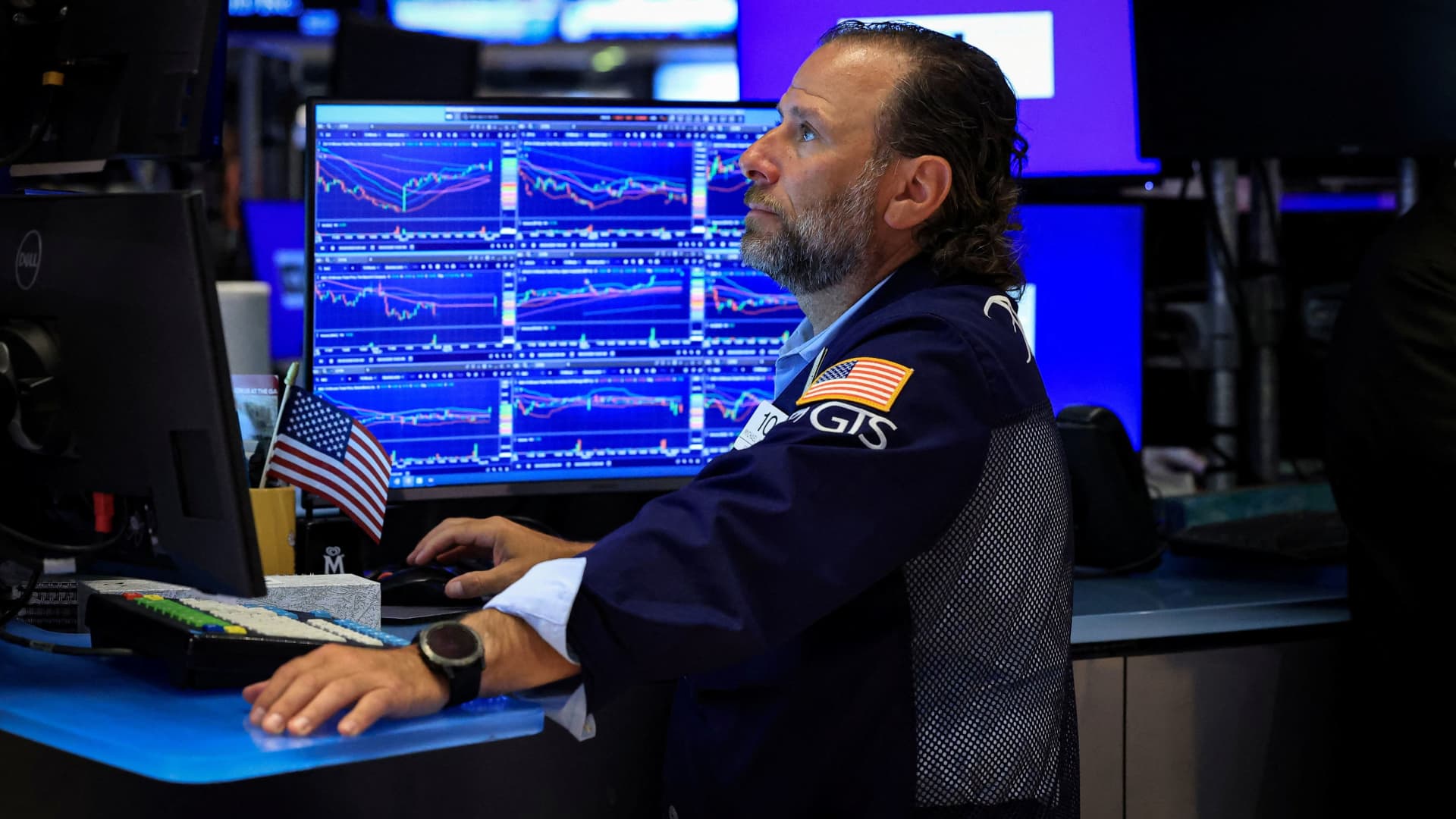 A trader works at a desk surrounded by blue financial market charts and U.S. flags.