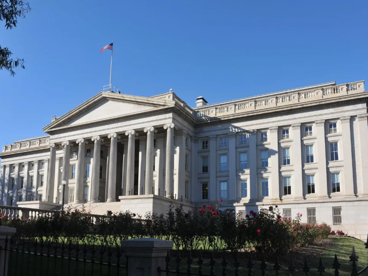 White neoclassical government building with columns, rose bushes, and an American flag under blue skies.