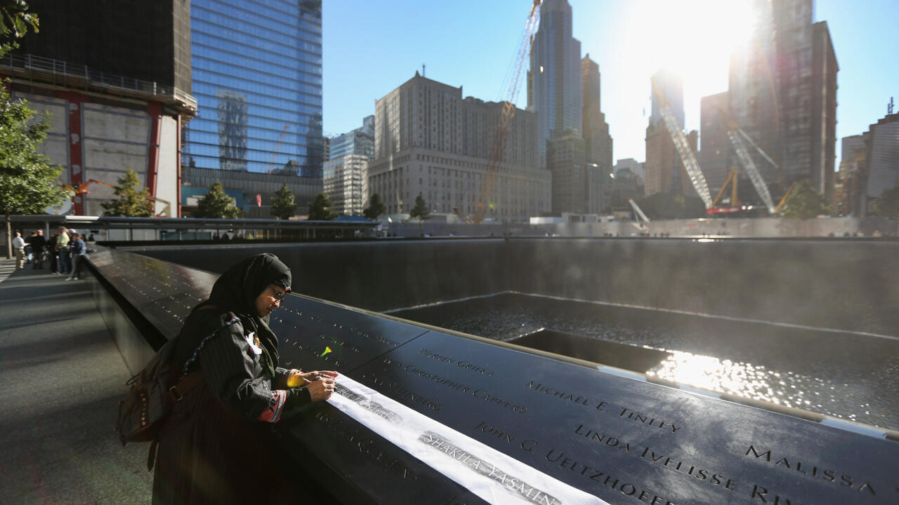 Person writes on a memorial parapet beside a reflecting pool amid sunlit skyscrapers.