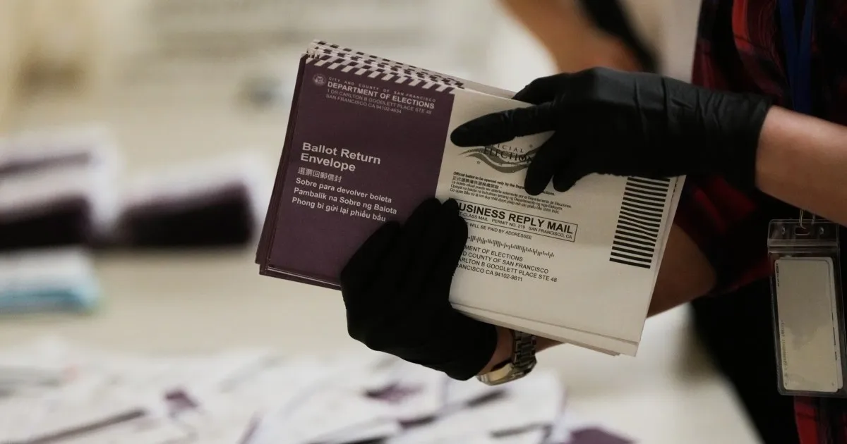 Gloved worker holds a stack of ballot return envelopes over a table.