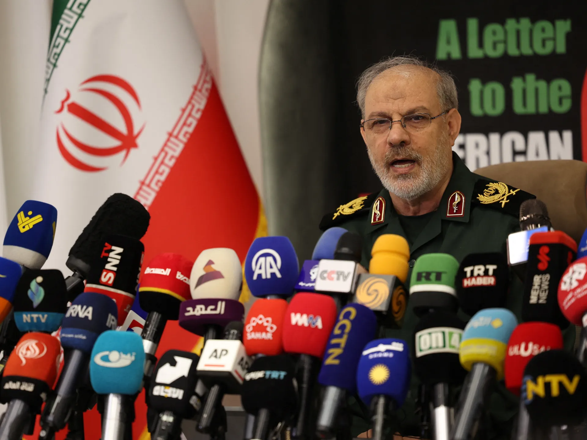 Iranian military official speaks at a press conference behind a dense cluster of microphones, with an Iranian flag behind him.
