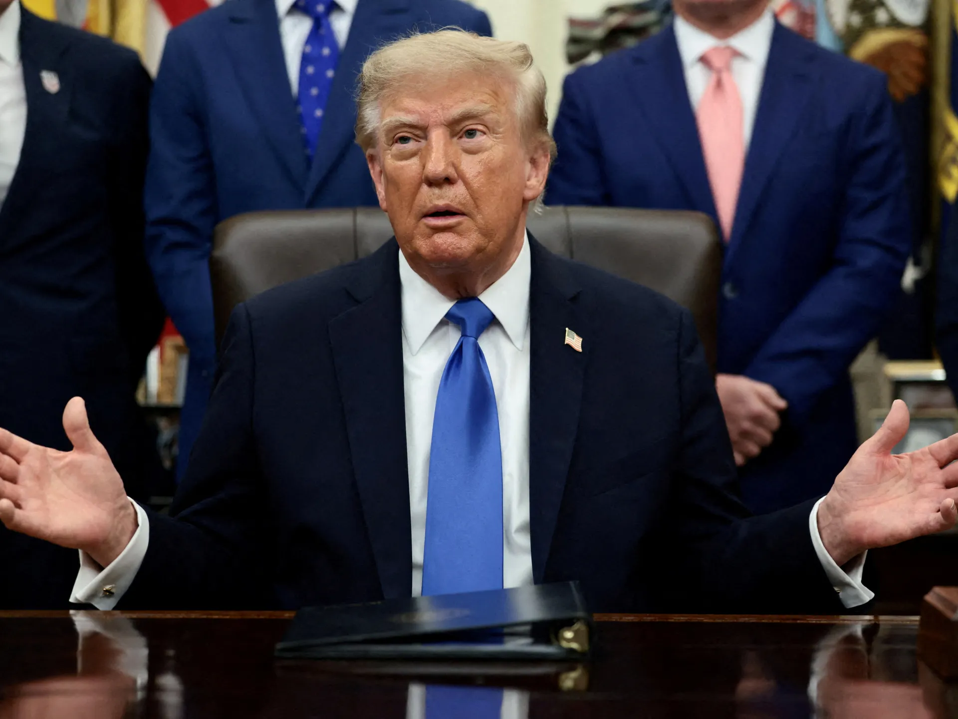 Donald Trump seated at a desk with both hands raised, officials standing behind him