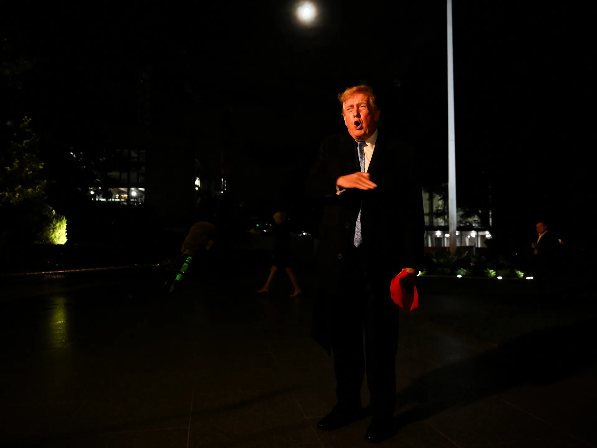 Donald Trump speaks outdoors at night, holding a red cap beside a microphone.