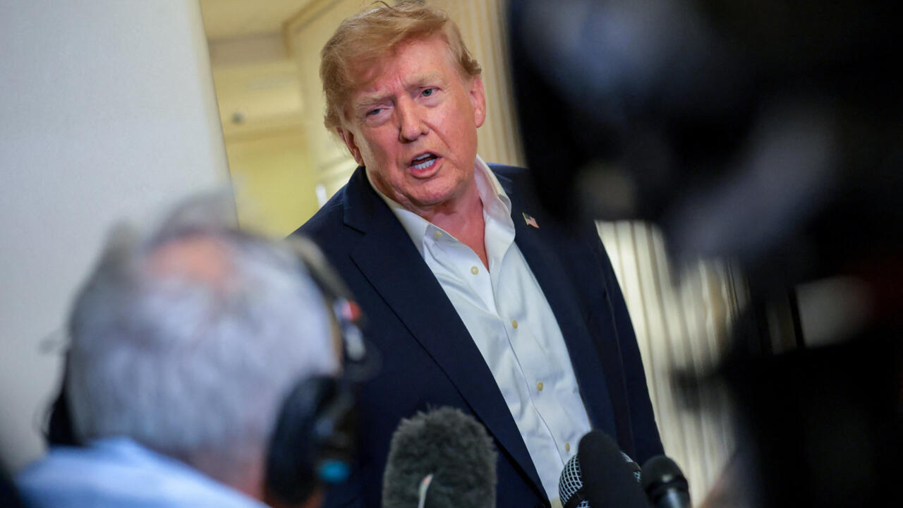 Donald Trump speaks to reporters while several microphones and cameras fill the foreground.