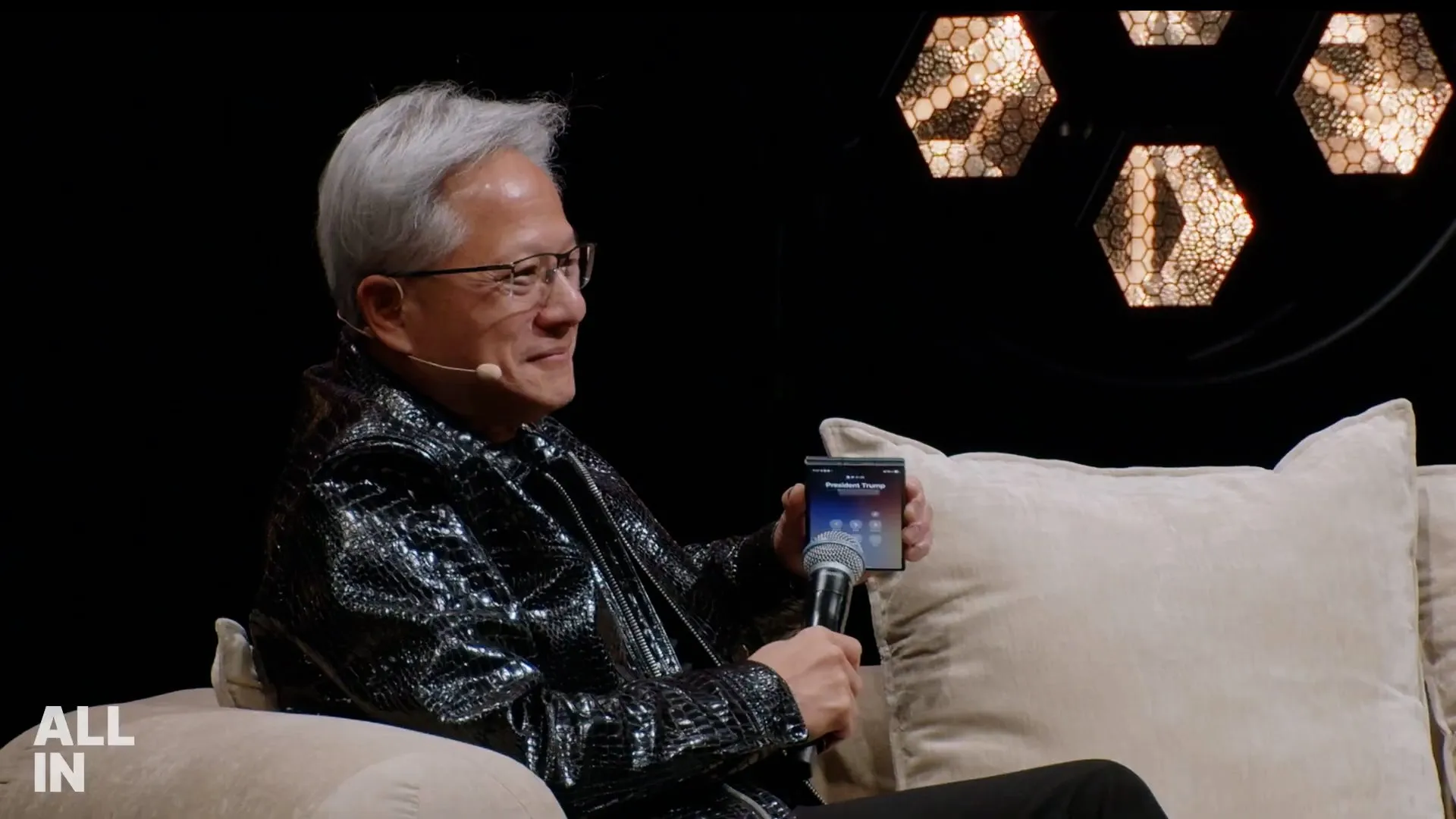Gray-haired man in glasses holds a smartphone and microphone while seated on a couch.