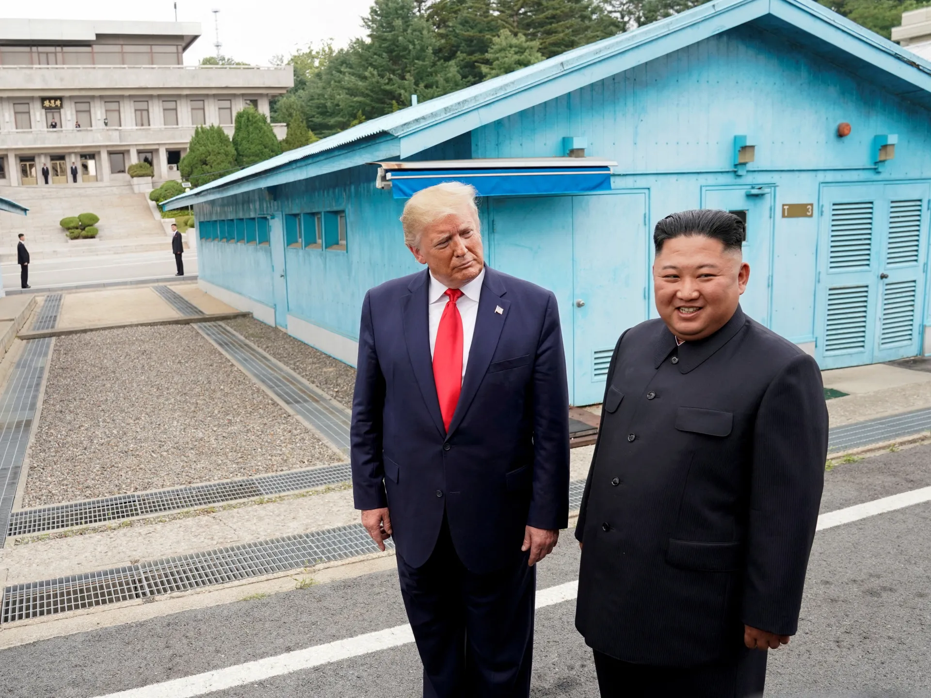 Donald Trump stands beside Kim Jong Un in front of a blue building, with guards and officials in the background.