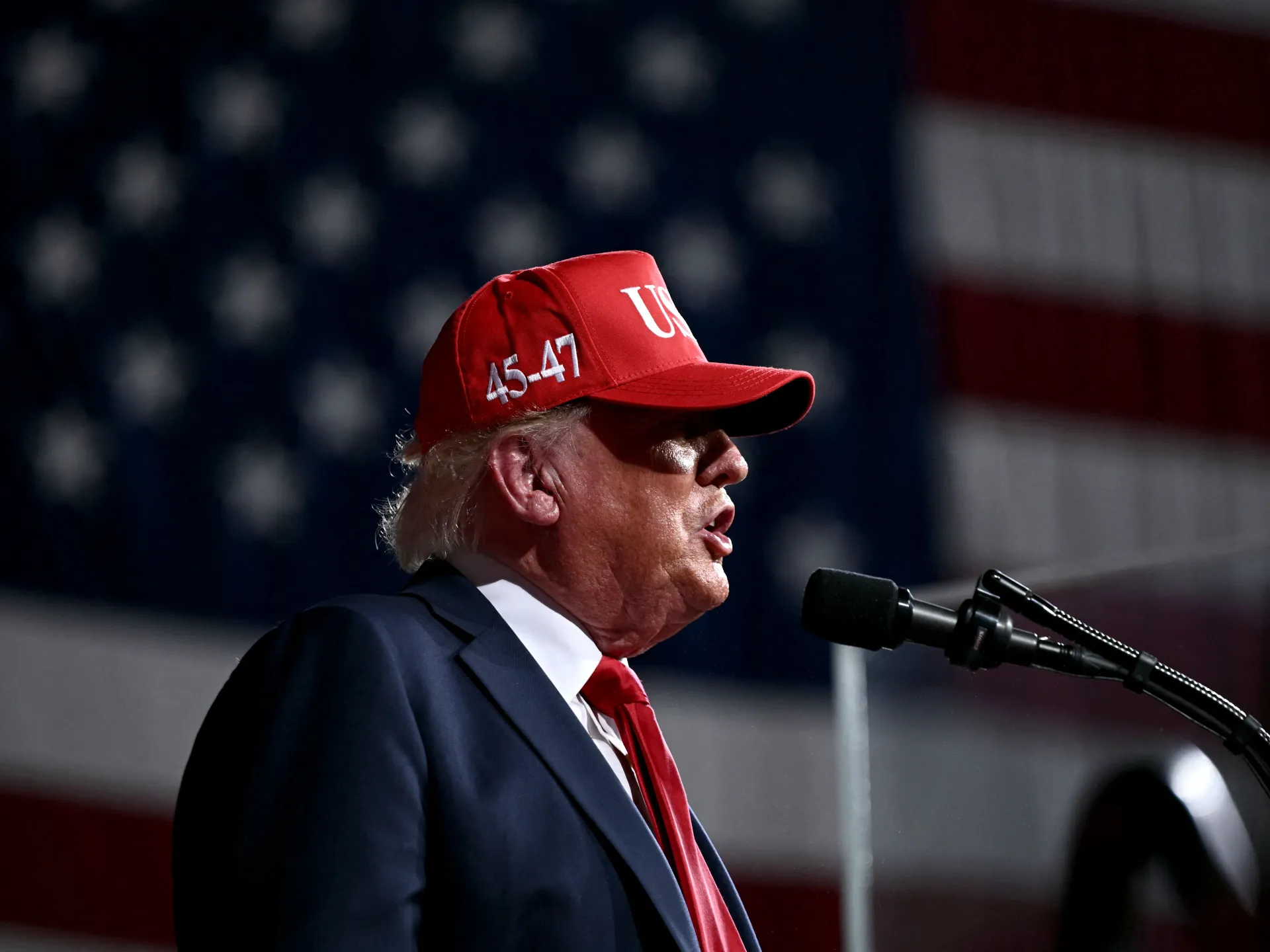 Donald Trump speaks at a podium wearing a red campaign cap and tie.