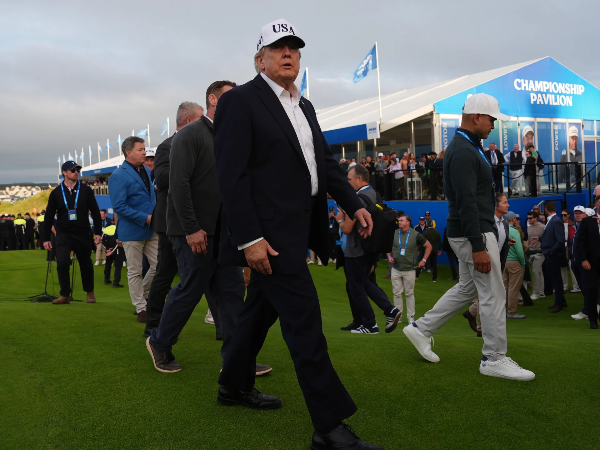 Man in dark suit and USA cap walks across a golf event beside spectators and officials.