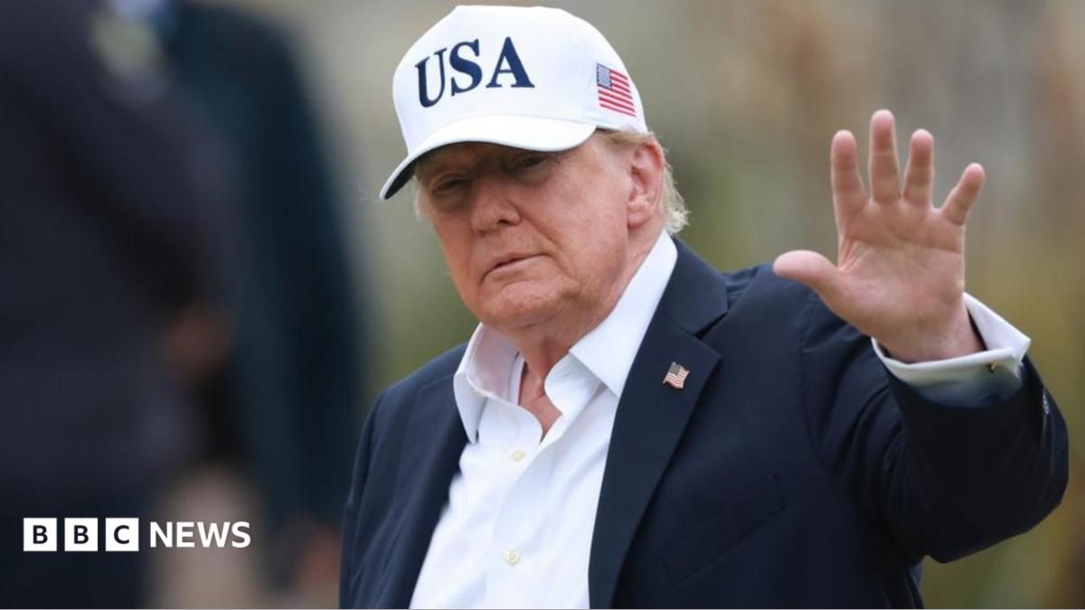 Donald Trump in a dark suit and USA cap raises his hand in a wave