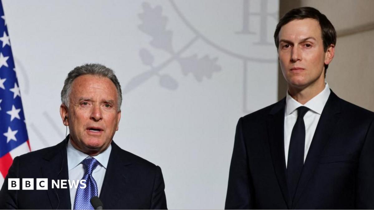 Two suited men stand at a press event beside a United States flag.