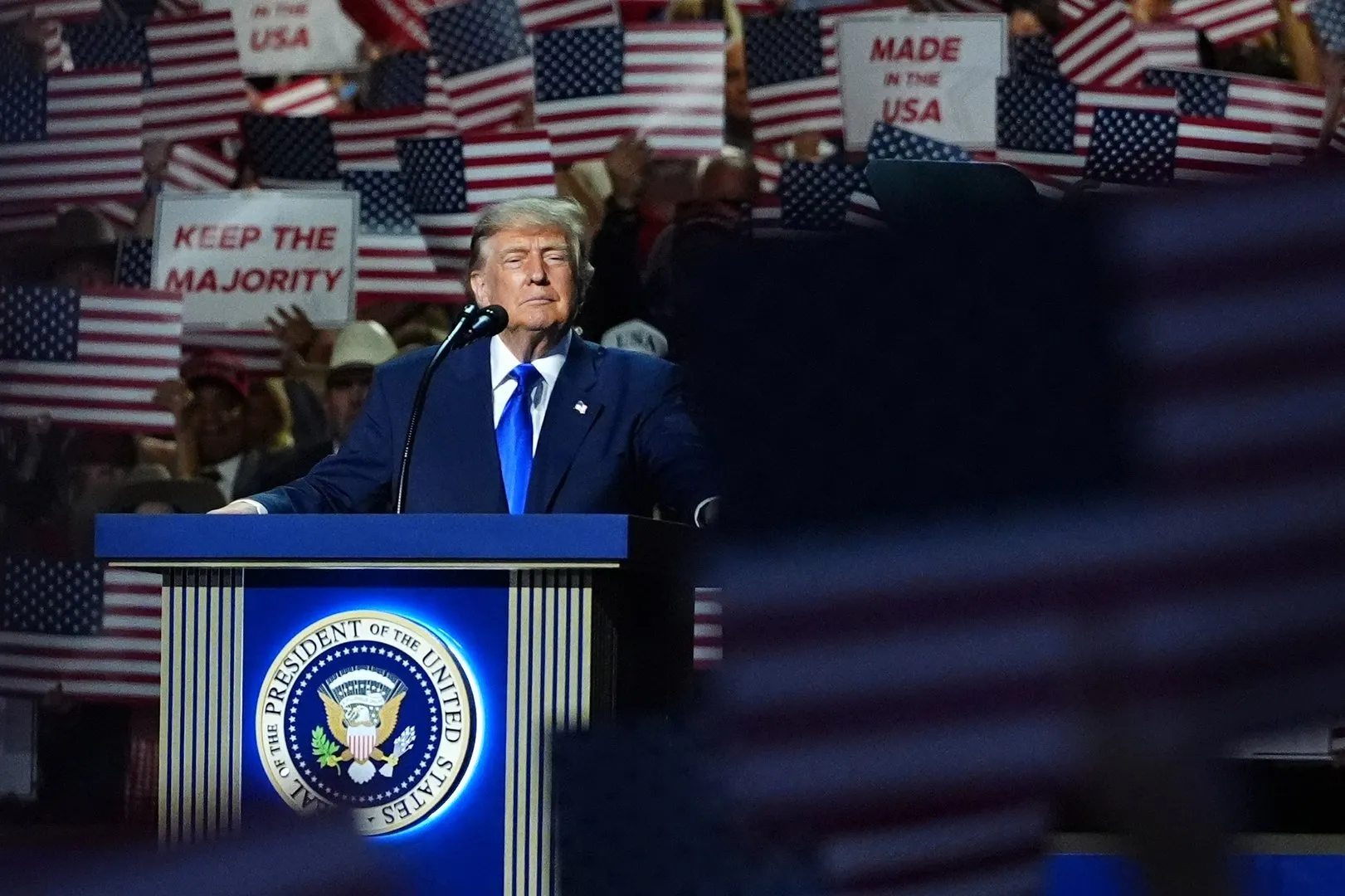 Donald Trump speaks at a podium before supporters holding American flags and campaign signs.