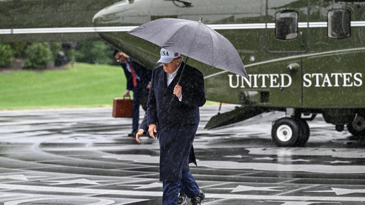 A man in a dark suit walks through rain holding an umbrella beside a U.S. aircraft.