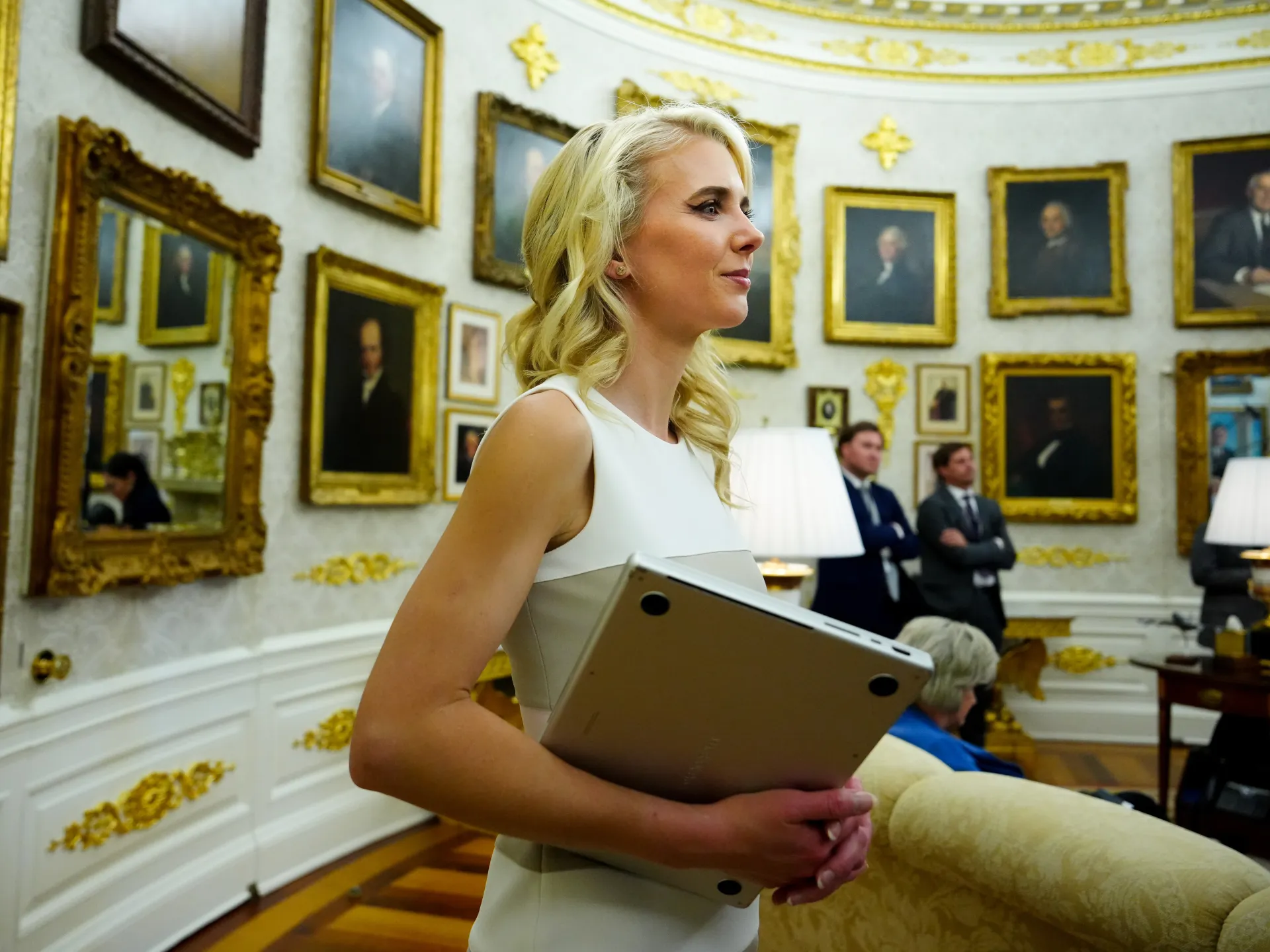 Blonde woman carrying a laptop through an ornate room lined with framed portraits and gold decorations.