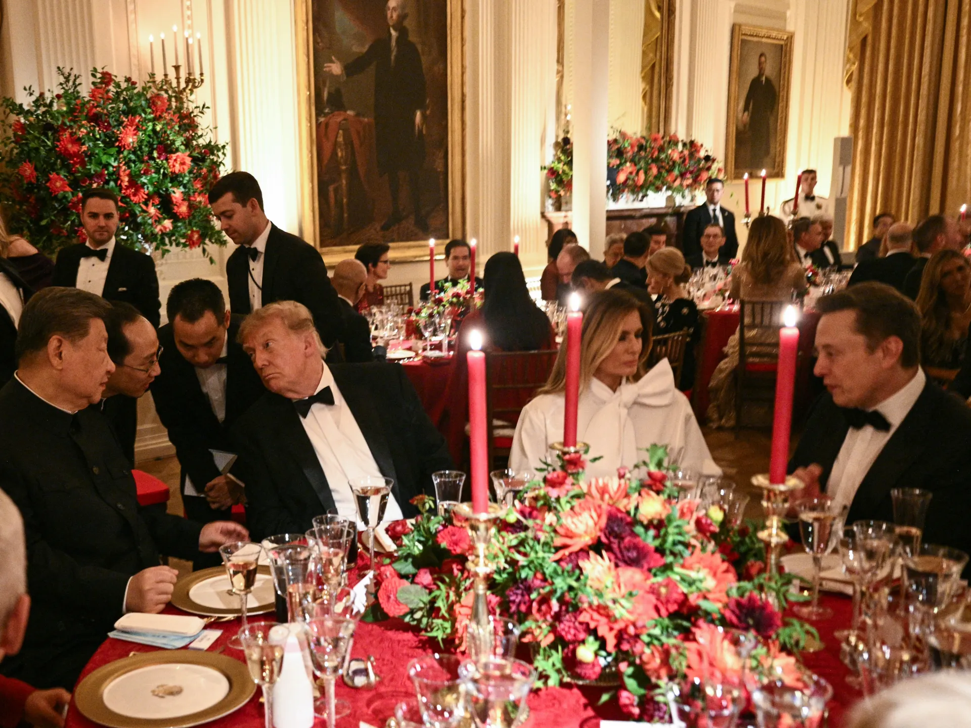 Guests in formal attire dine at a candlelit banquet table with a large floral arrangement.