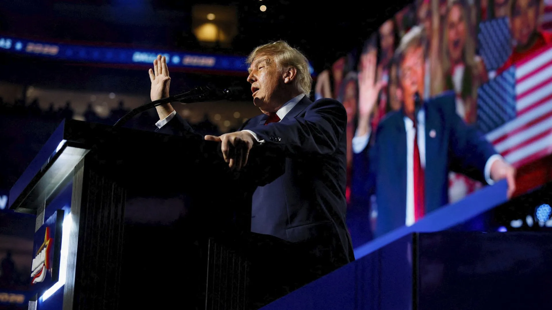 Donald Trump speaks at a podium, raising one hand and pointing toward the audience.