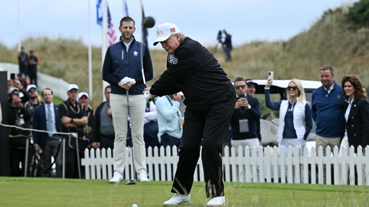 Older man in black golf attire swings a club as spectators watch behind a white fence.