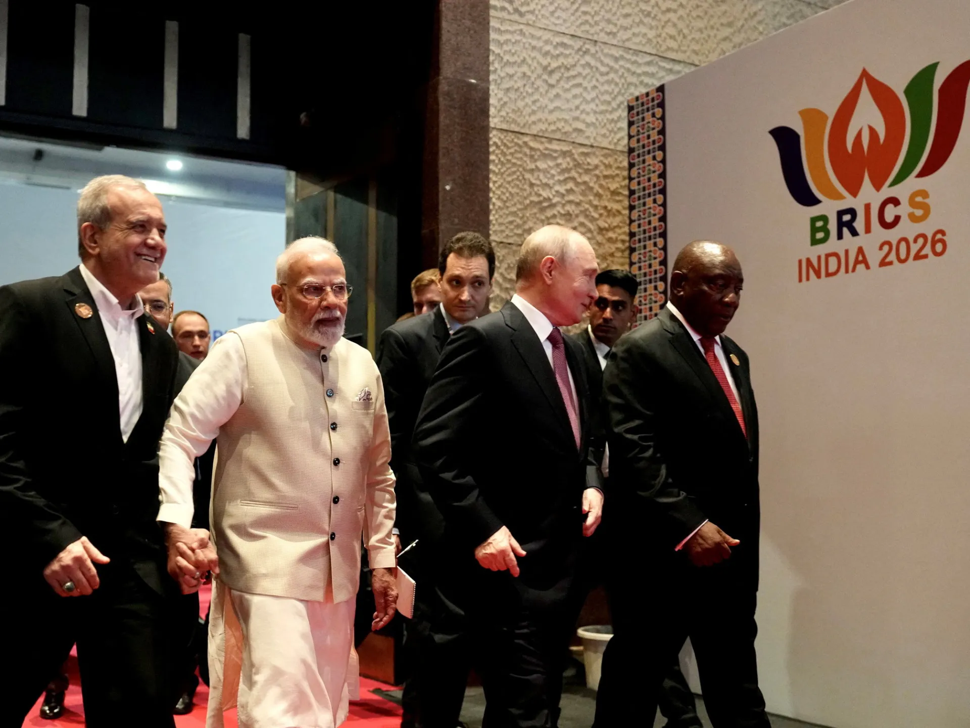 Several political leaders walk together past a BRICS India 2026 conference sign.