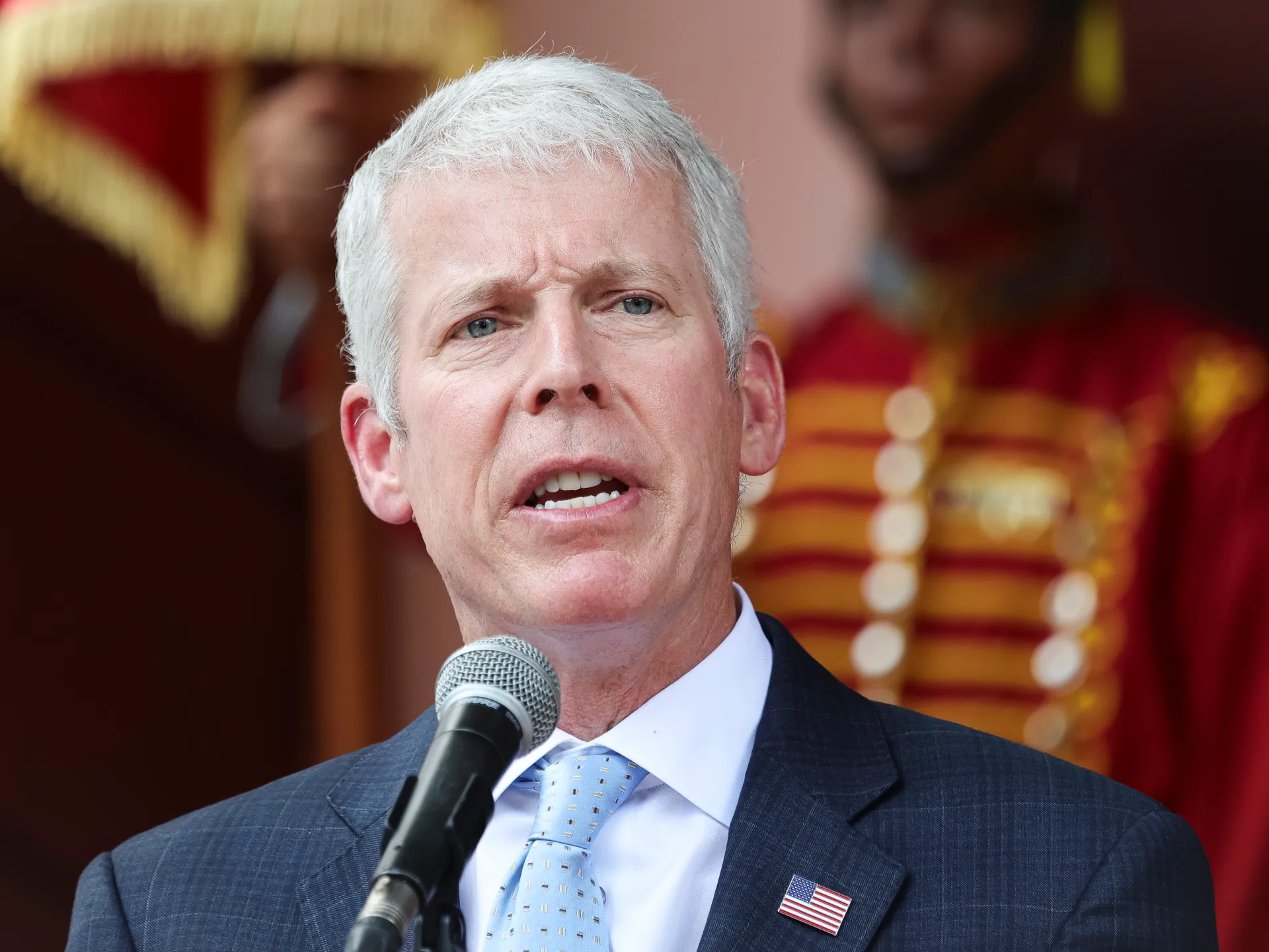 White-haired man in a suit speaking into a microphone with a U.S. flag lapel pin