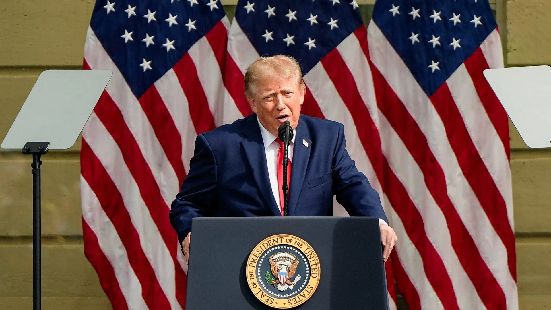 Donald Trump speaks at a podium surrounded by American flags and teleprompter screens.