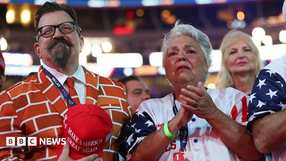 Three spectators watch an event, including a man in an orange patterned jacket and women in patriotic clothing.