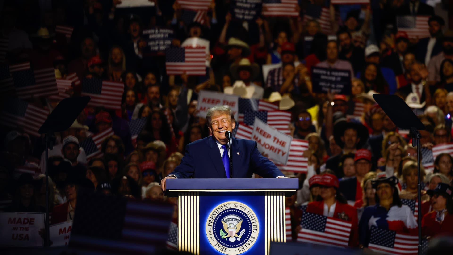Donald Trump speaks at a podium before a crowd holding American flags and campaign signs.