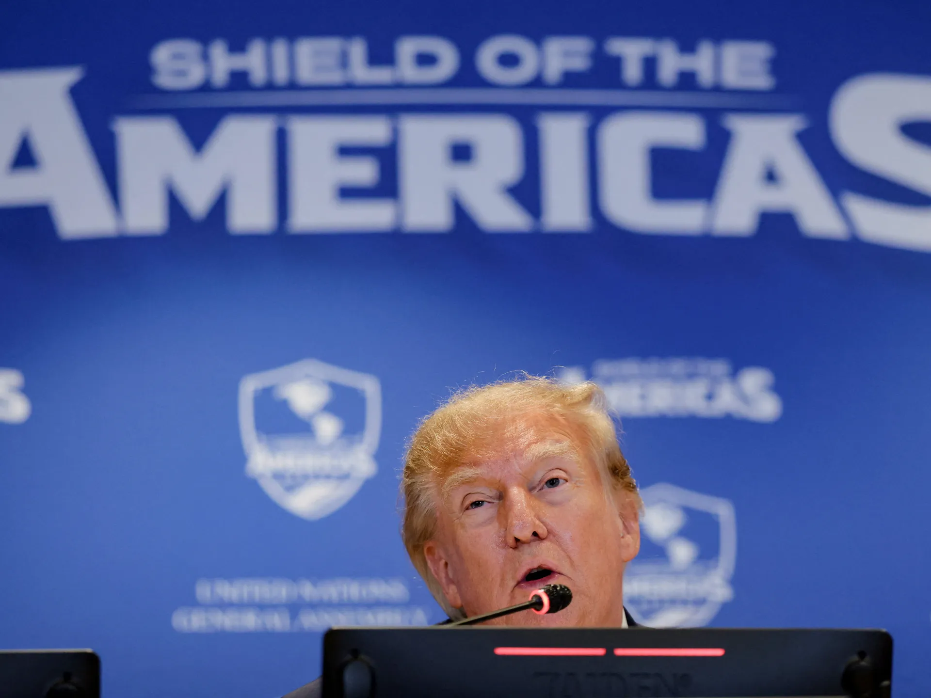 Donald Trump speaks into a microphone at a podium against a blue Shield of the Americas backdrop.
