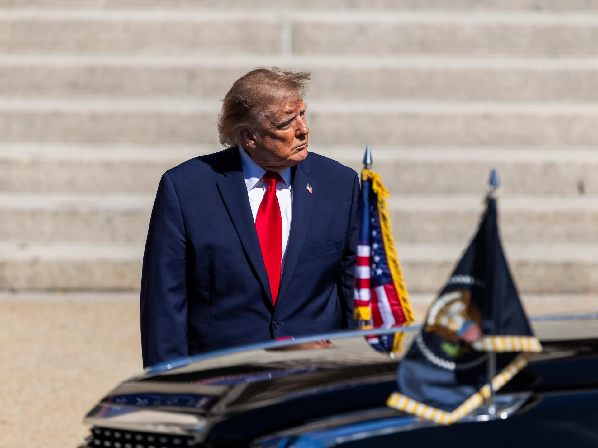 Donald Trump in a dark suit and red tie stands beside a car and flags.