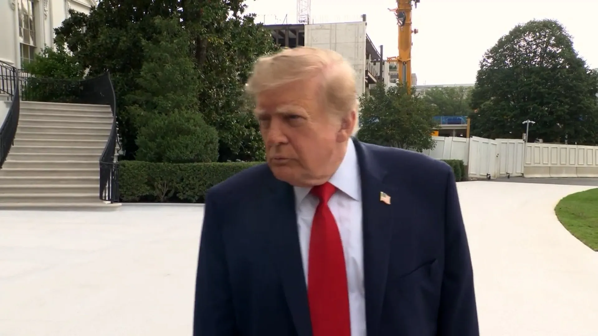Donald Trump in a navy suit and red tie stands outdoors near a staircase and construction crane.