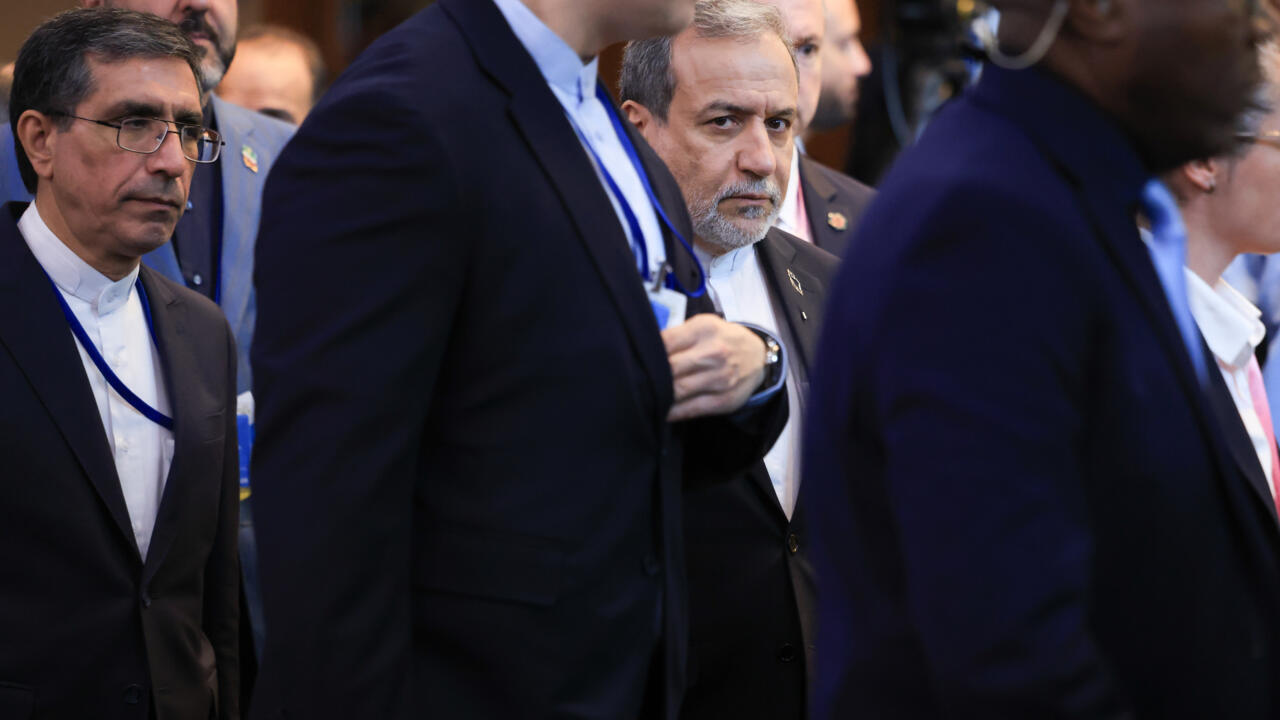 Men in suits and lanyards stand in a crowded indoor gathering, with one man partly obscured in the foreground.