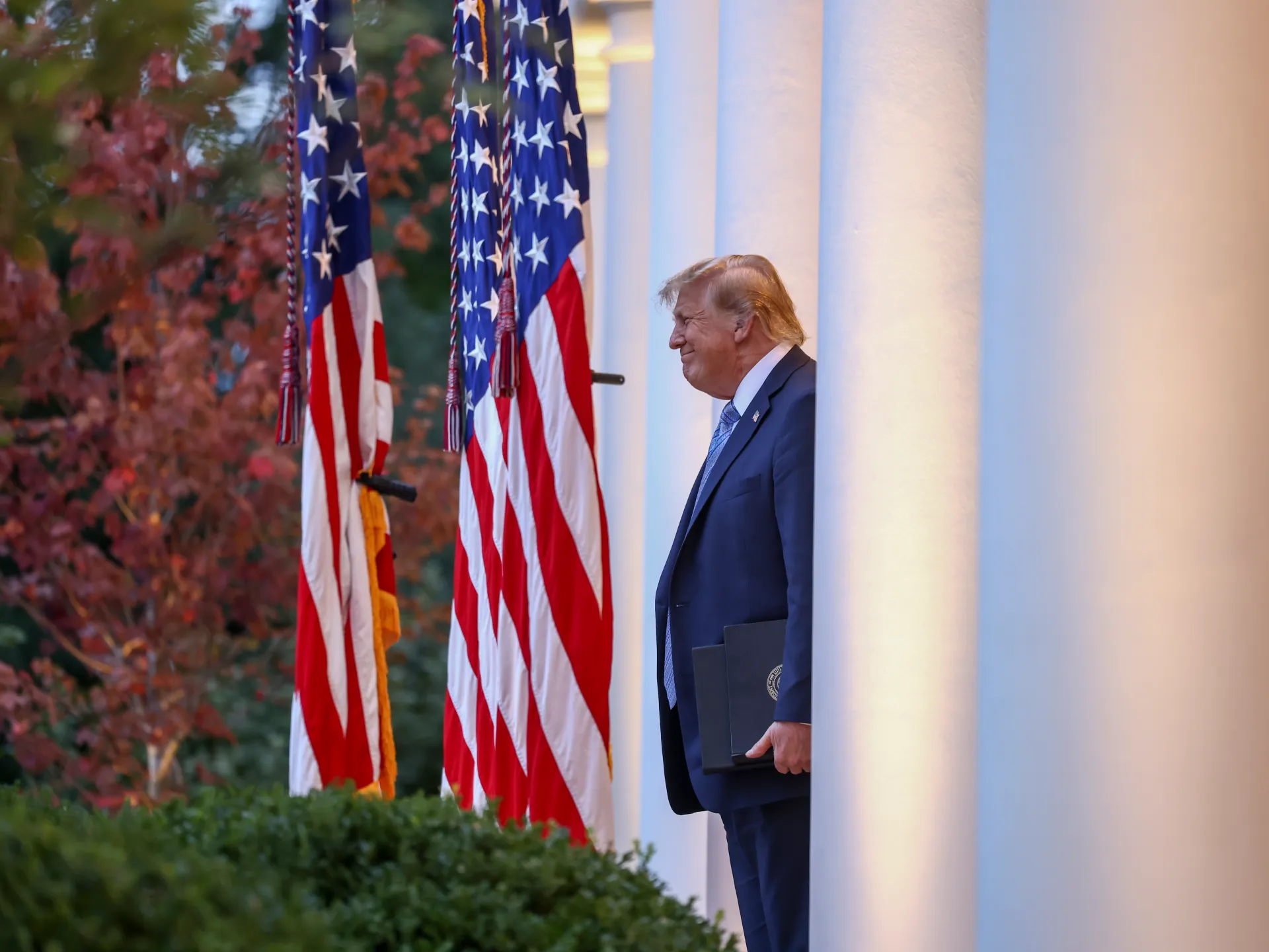 Donald Trump stands beside American flags near a column, holding a dark folder.