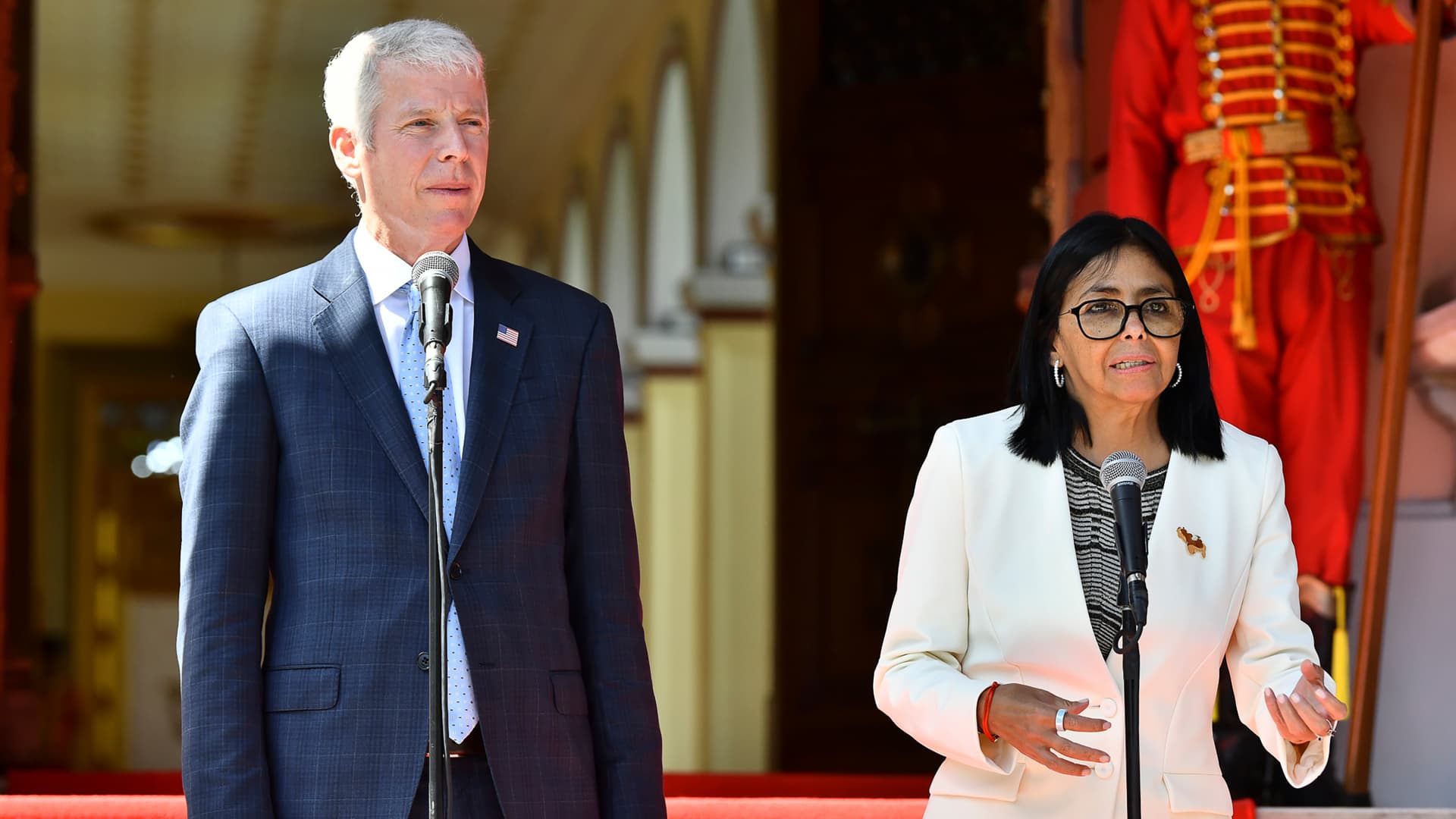 Two officials speak at microphones outside a building, with a ceremonial guard behind them.