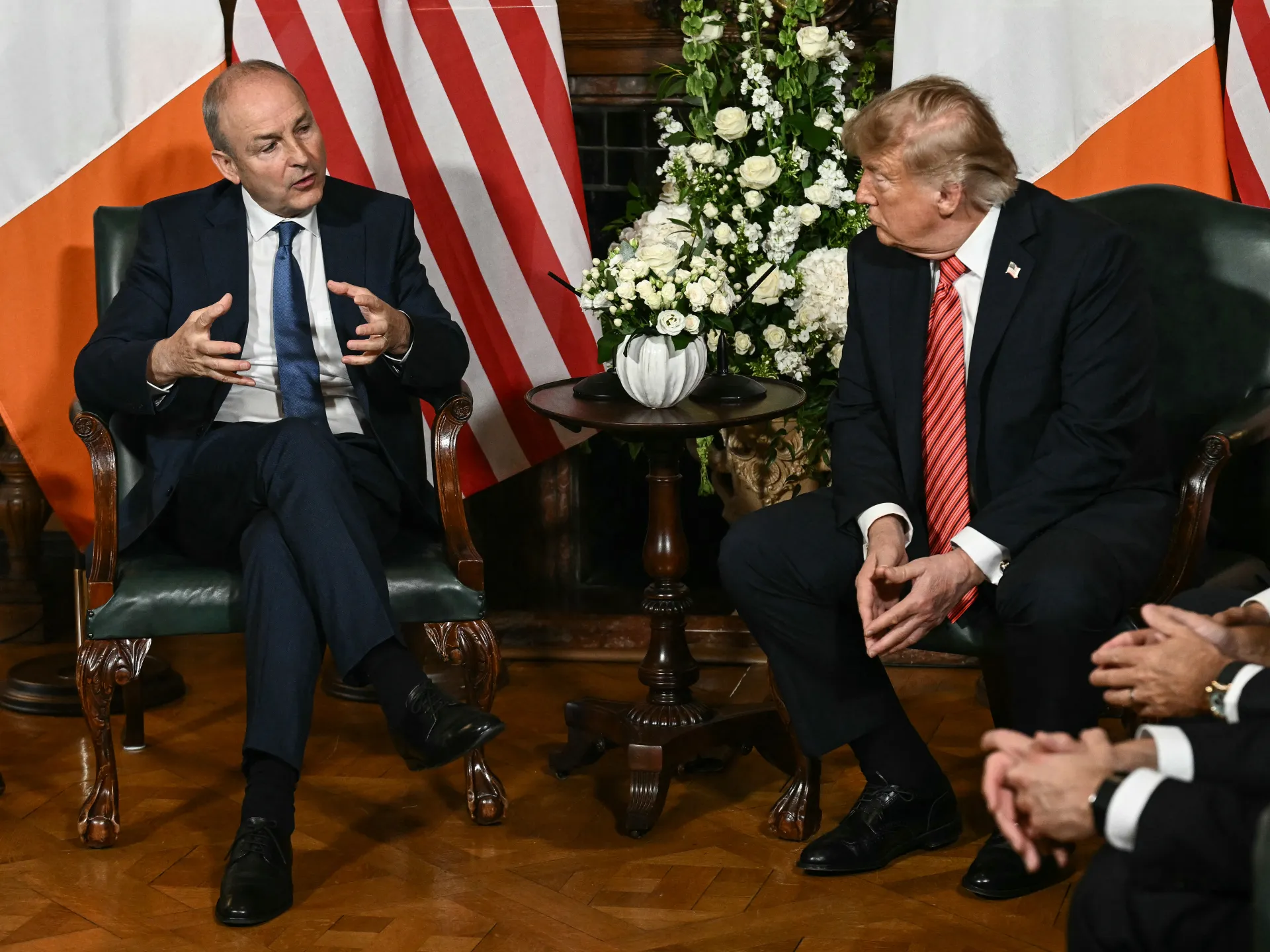 Two suited men sit in armchairs conversing beside floral arrangements and American and Irish flags.