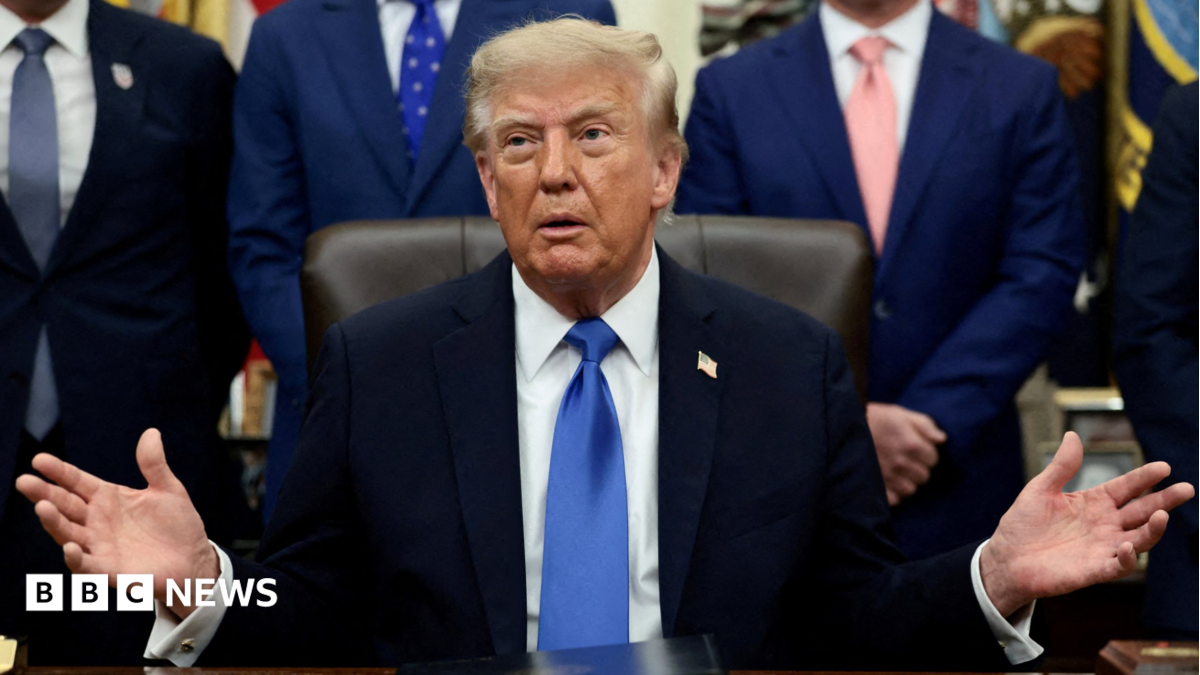 Donald Trump sits in a chair with both hands raised, surrounded by suited officials.