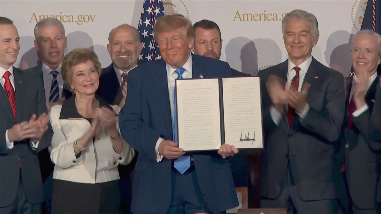 Donald Trump holds an open signed document as officials stand beside him and applaud.