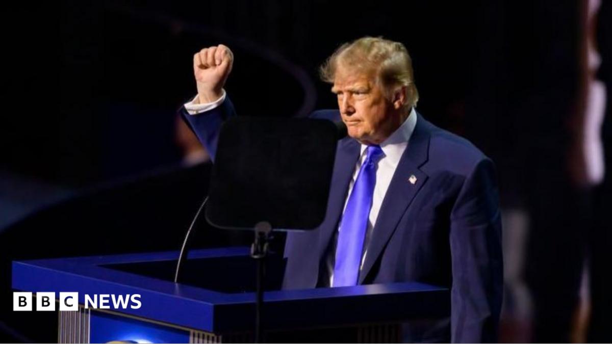Donald Trump raises a fist while standing at a podium behind a teleprompter.